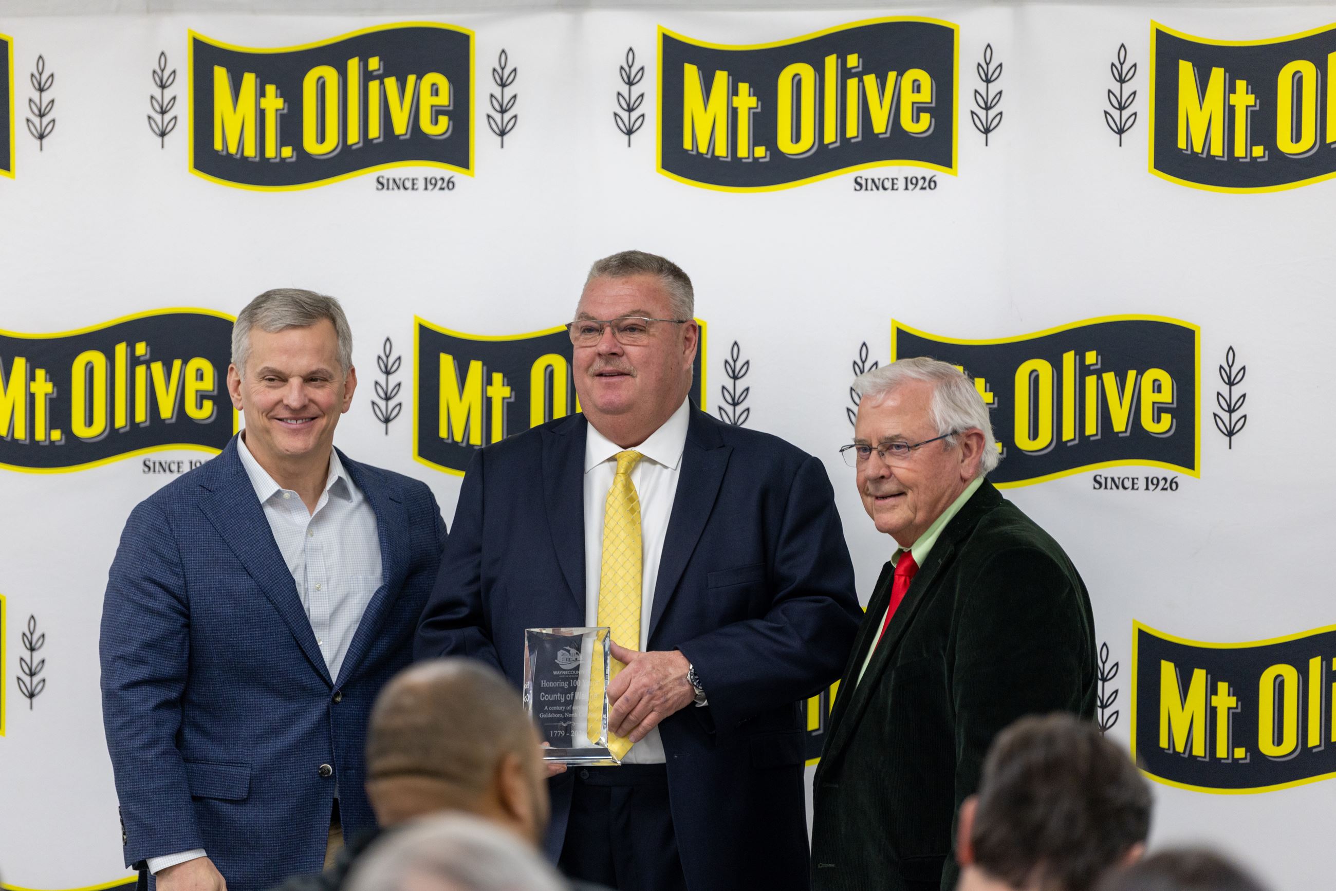 Three men at an award ceremony in front of a Mt. Olive backdrop.